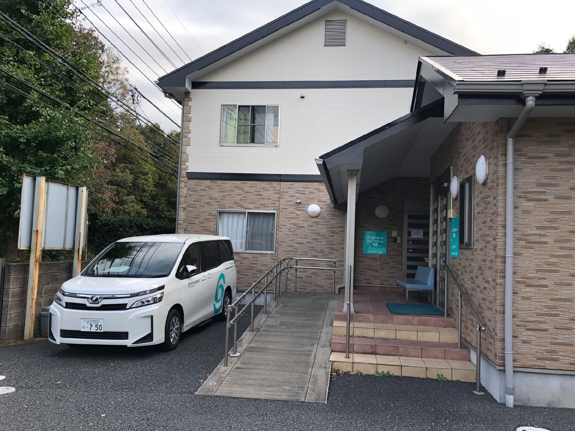 駐車場にはホームの福祉車両が止まっていて、レンガ調の壁に三角屋根が付いた建物がある。