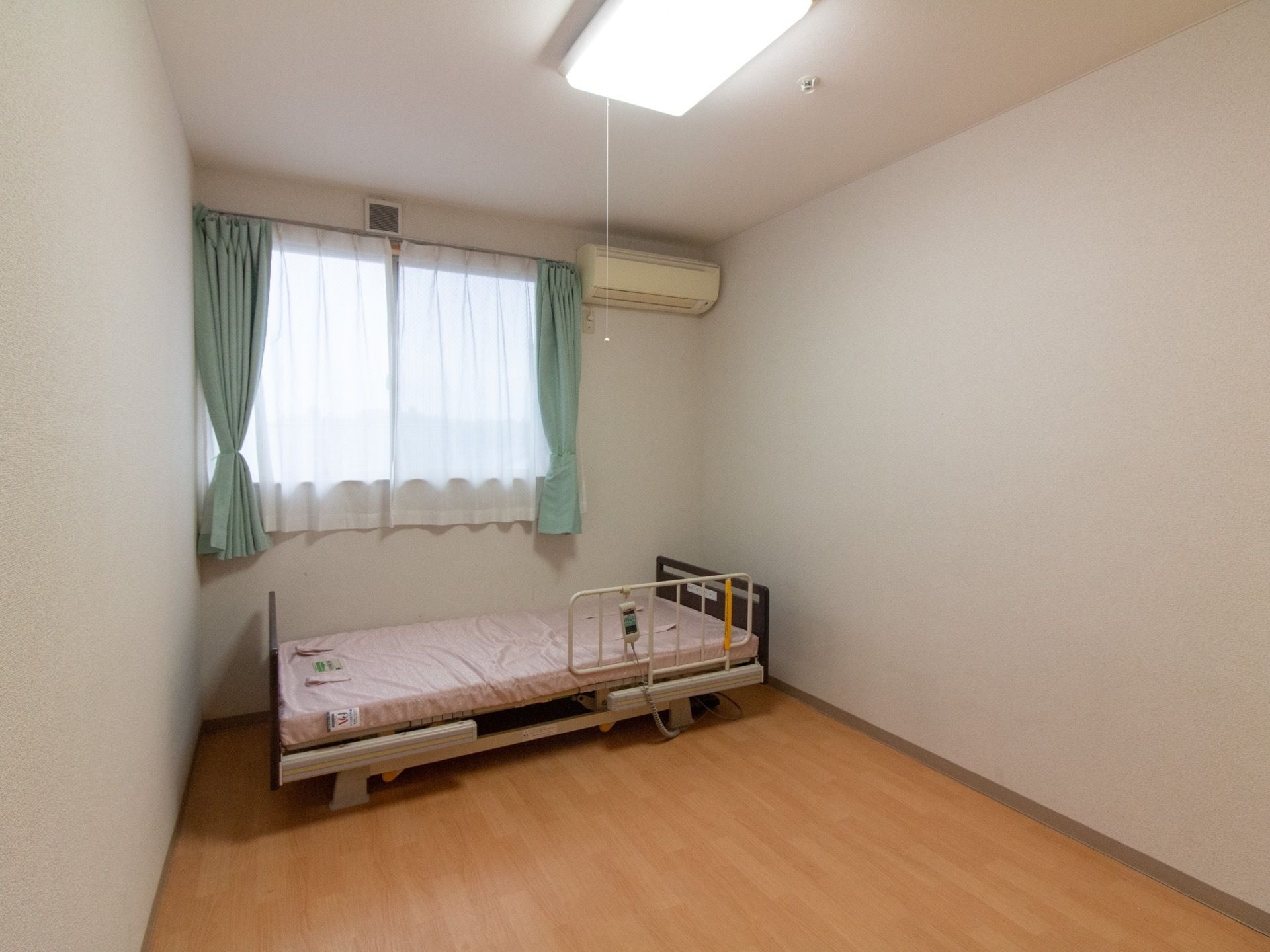 フローリングの床で、窓にはカーテンがかかっている。窓の手前にベッドがあり、壁にはエアコンが設置されている。