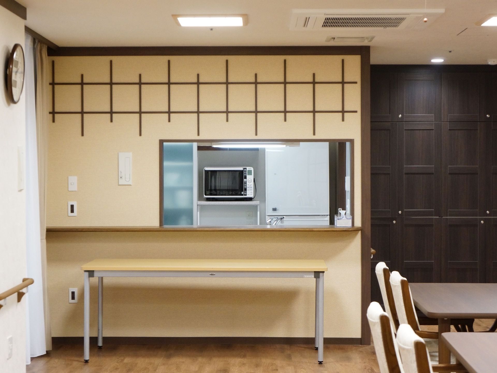ダークブラウン、ベージュを基調としたシックな食堂。カウンター奥には電子レンジが見える。右手にはテーブル、左手には時計が壁にかかっている。
