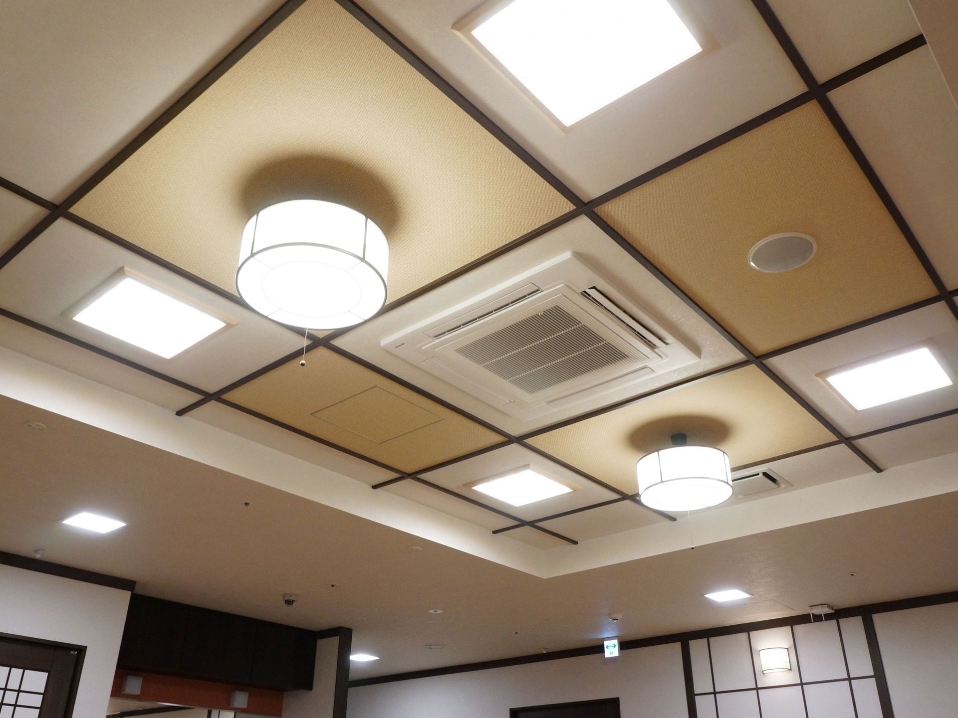 食堂の天井が写っている。正方形と円形の照明があり、天井のデザインはベージュと白色がタイル状に並んでいる。中心には空調が設置されている。