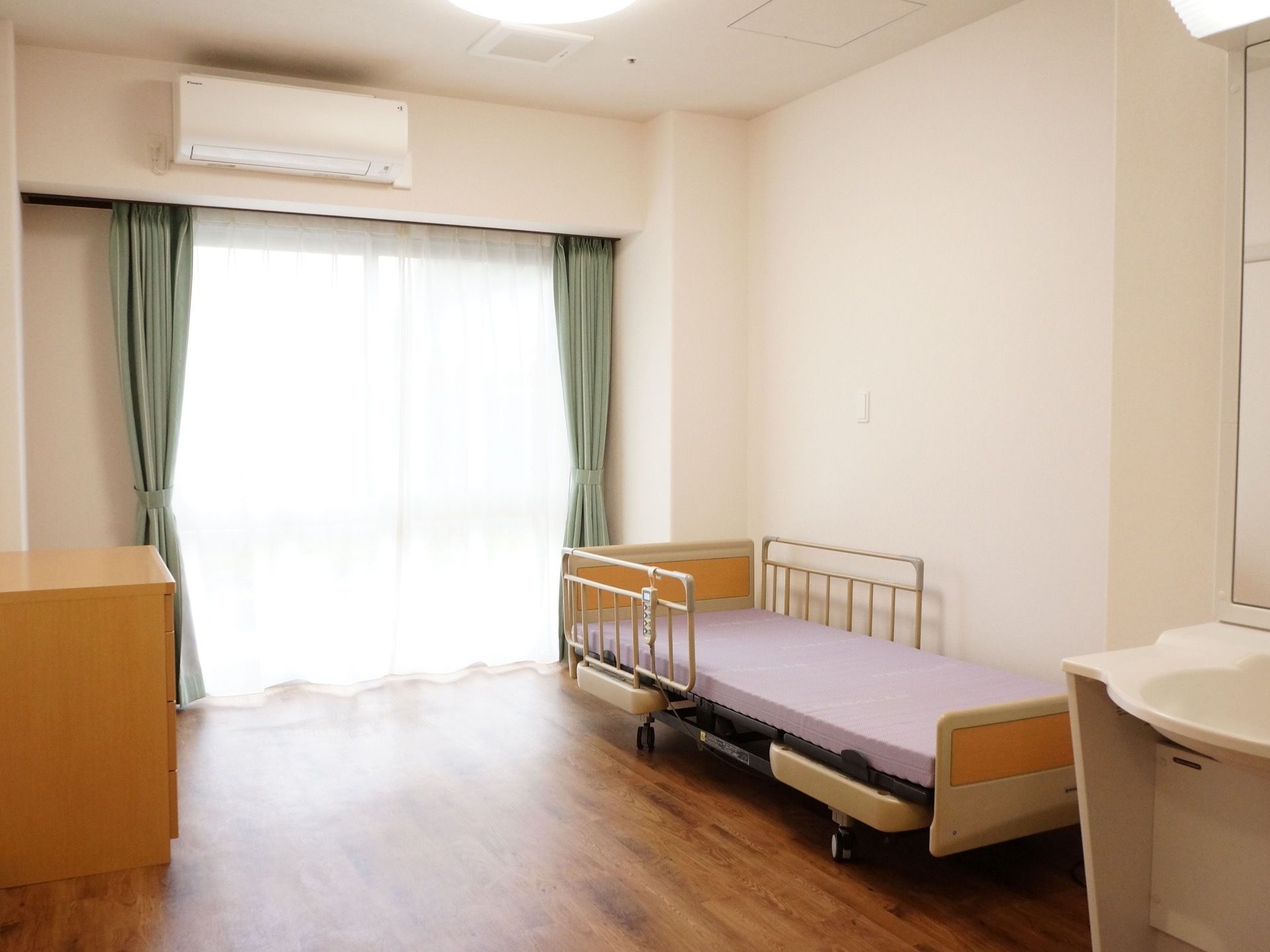 居室の正面には大きなガラス戸があり、壁の上部にはエアコンが取り付けられている。また、壁際には介護用ベッドが備え付けられ、向い側には棚がある。