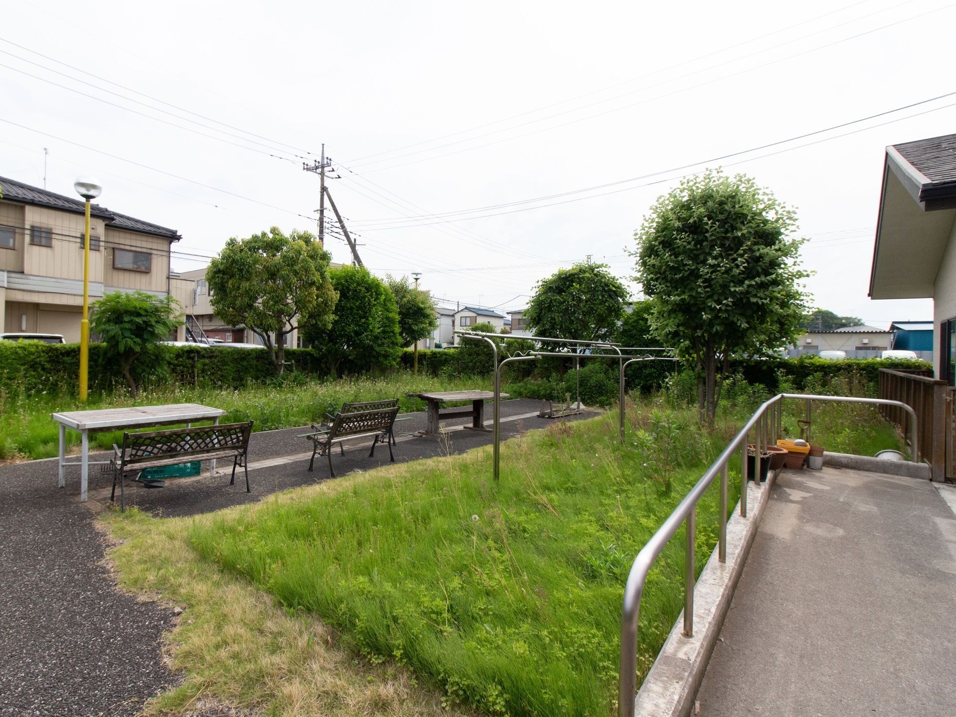 舗装された通路に机が２卓、椅子が３脚置かれている。スロープとベンチの間の草地には、物干し台が設置されている。