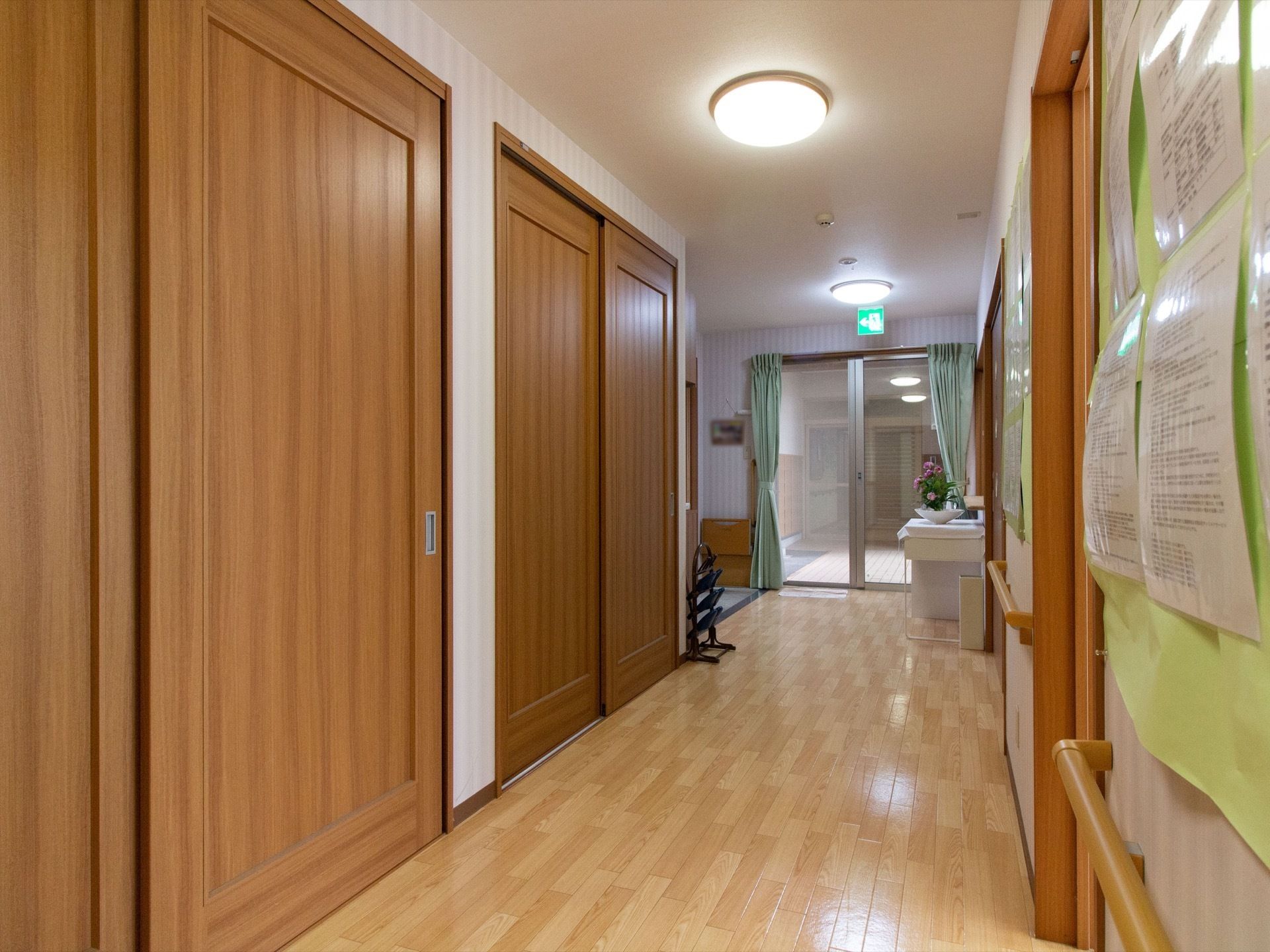 居室エリアの廊下である。居室の扉が並び、片面の壁には手すりが設置されている。手すりの上の壁に掲示物が貼られている。