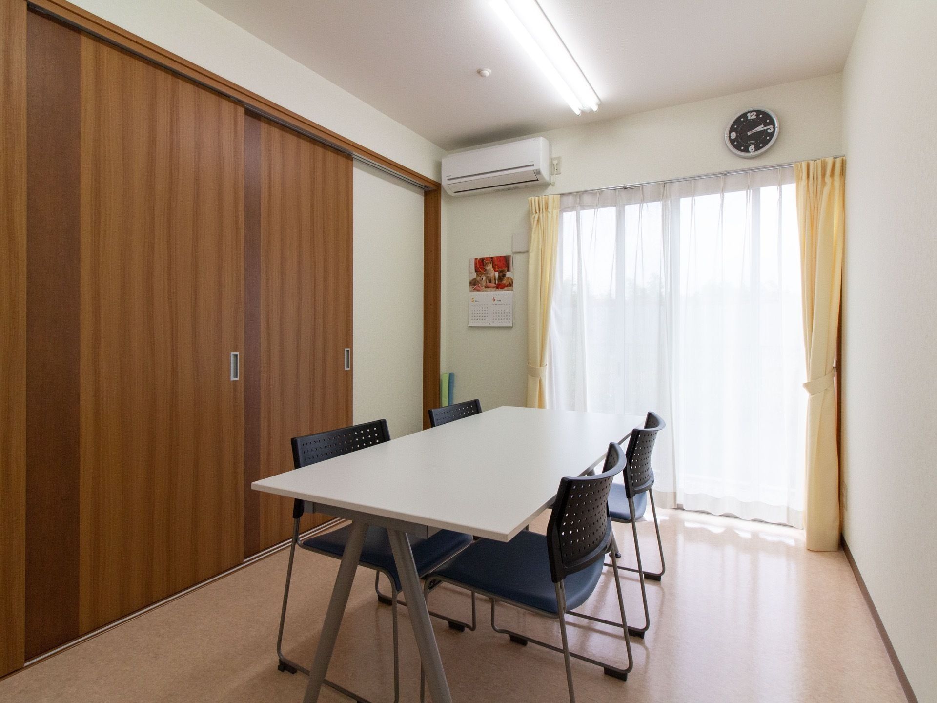 部屋の中央に、白いテーブルが置かれている。テーブルには４脚の椅子が片面２脚ずつ配置されている。部屋にはエアコンが設置されている。