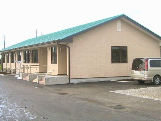 玄関に手すり付きのスロープが付き、緑の三角屋根が付いた日当たりのいい平屋建ての建物。駐車場には車も止まっている。