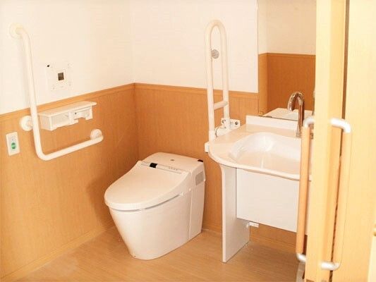 エル字型の手すりや可動式の手すりが付いた広いトイレ。手洗い場もあり、オレンジの呼び出しボタンも付いている。