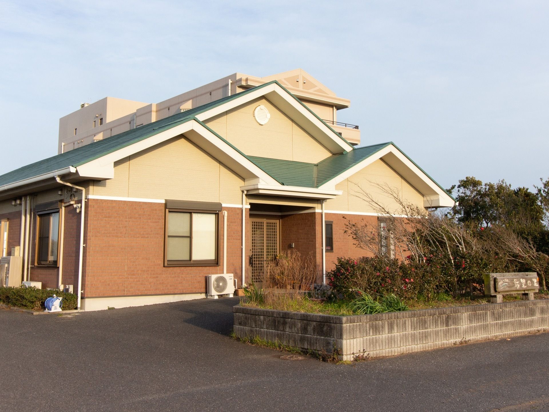 クリーム色とブラウンのツートンカラーの外壁、緑の三角屋根の１階建ての外観である。敷地内には、豊富な植栽を取り入れている。