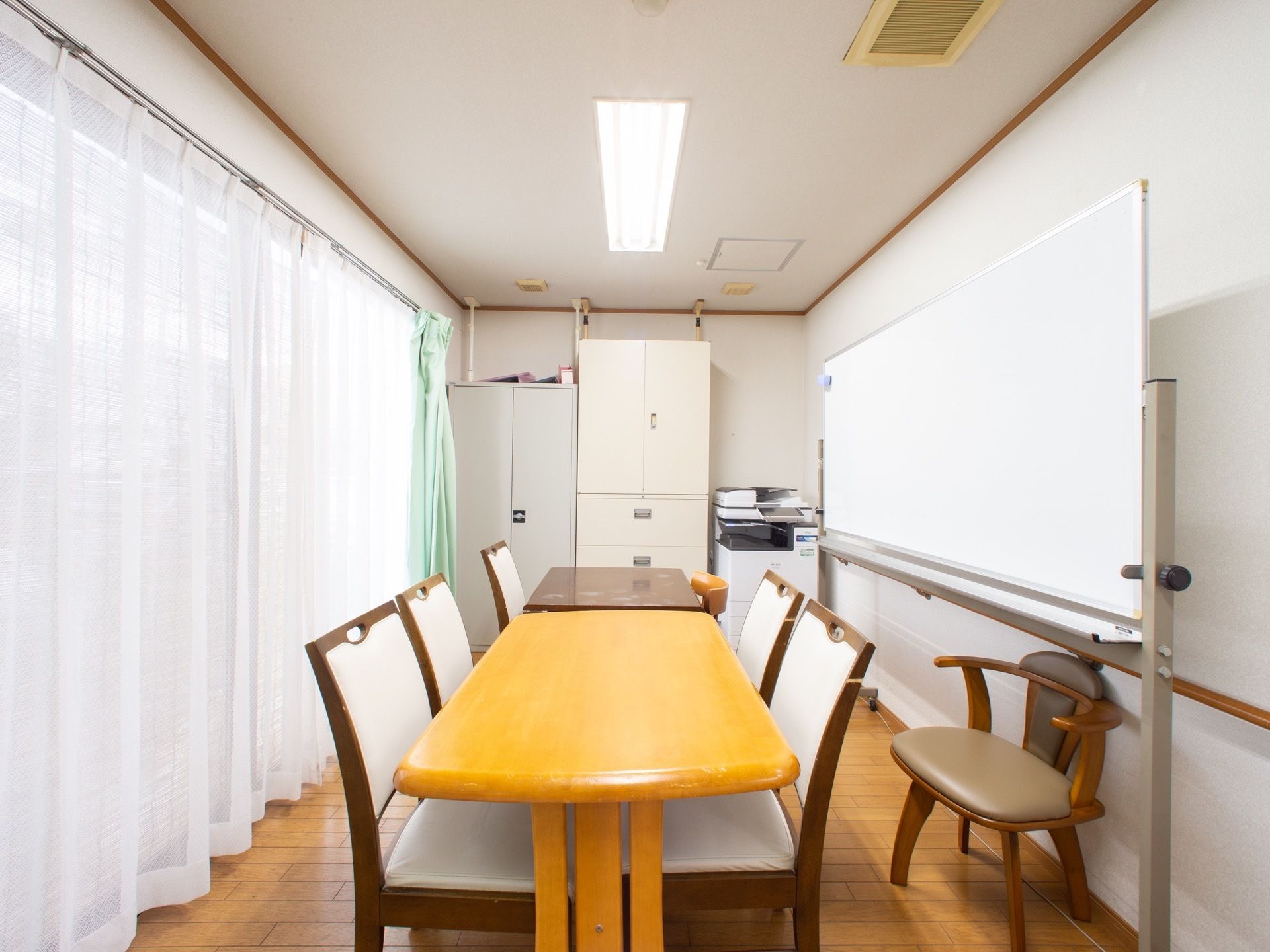 相談室は奥行きがあり、長机と椅子が７脚用意されている。奥には、収納棚２台とプリンターがある。