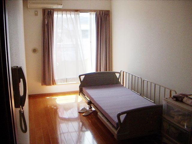レースカーテンとカーテンが付いた窓があるフローリングの部屋にはエアコンや介護ベッドが設置されている。