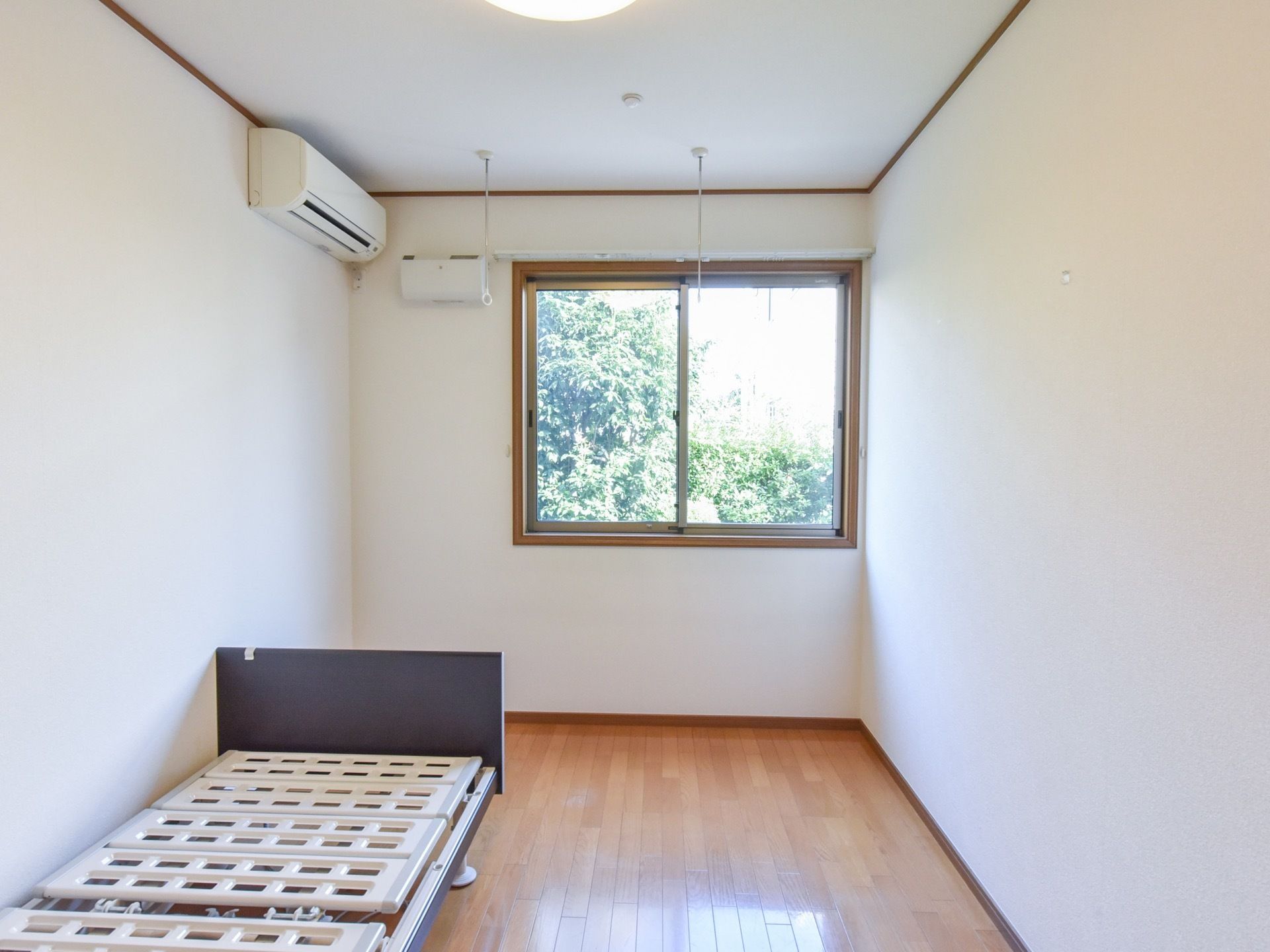 居室には低宋のベッドが用意されている。床はライトブラウンのフローリングで、窓からは緑が見える。エアコンが設置されている。