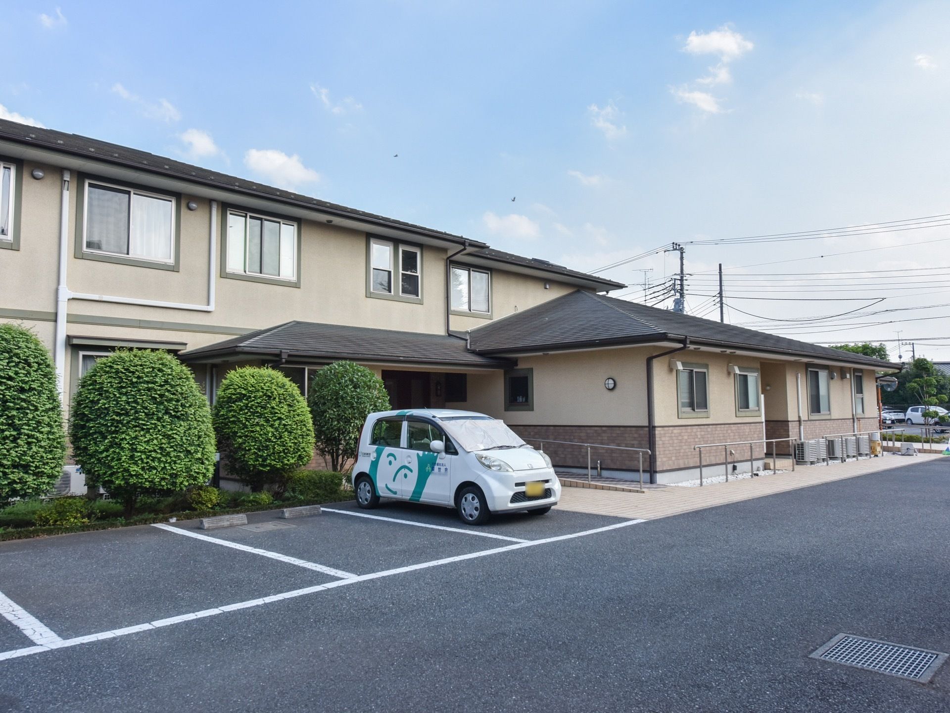施設の前に舗装された駐車場があり、施設の車が停められている。建物と駐車場の間には手入れのされた植木がある。