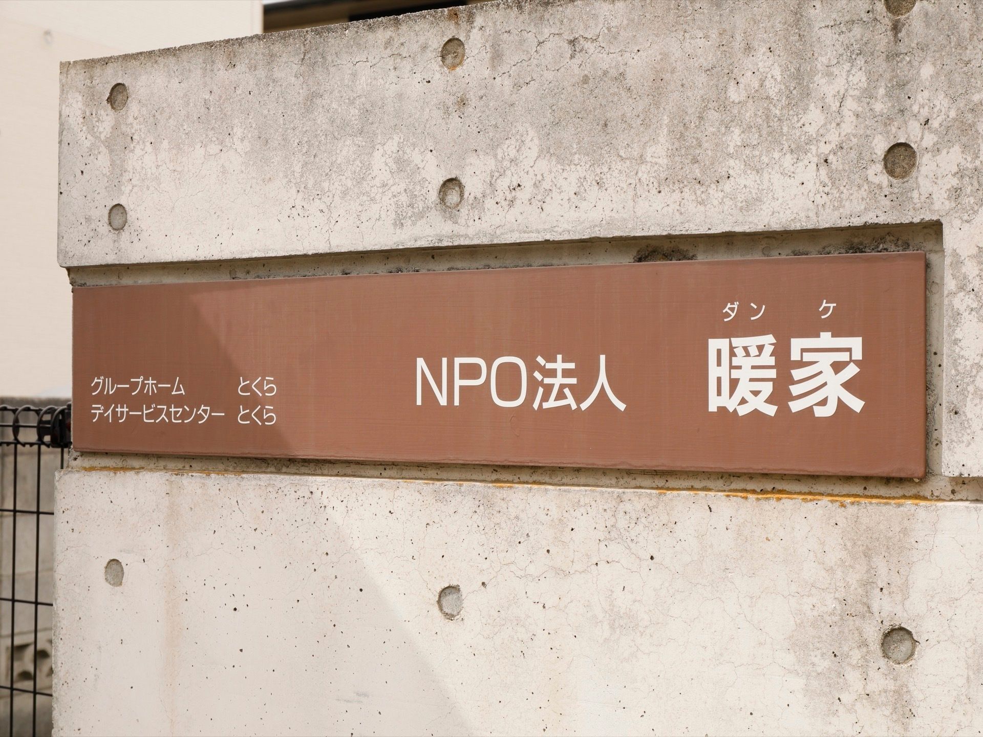 敷地出入り口のコンクリート塀には、施設名が記された茶色い長方形の看板が設置されている。左横には、黒いフェンスが取り付けられている。