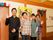 年代の違う４人のスタッフが笑顔で横一列に並んで立っている。背後の壁には手すりが設置されており、大きな絵が飾られている。