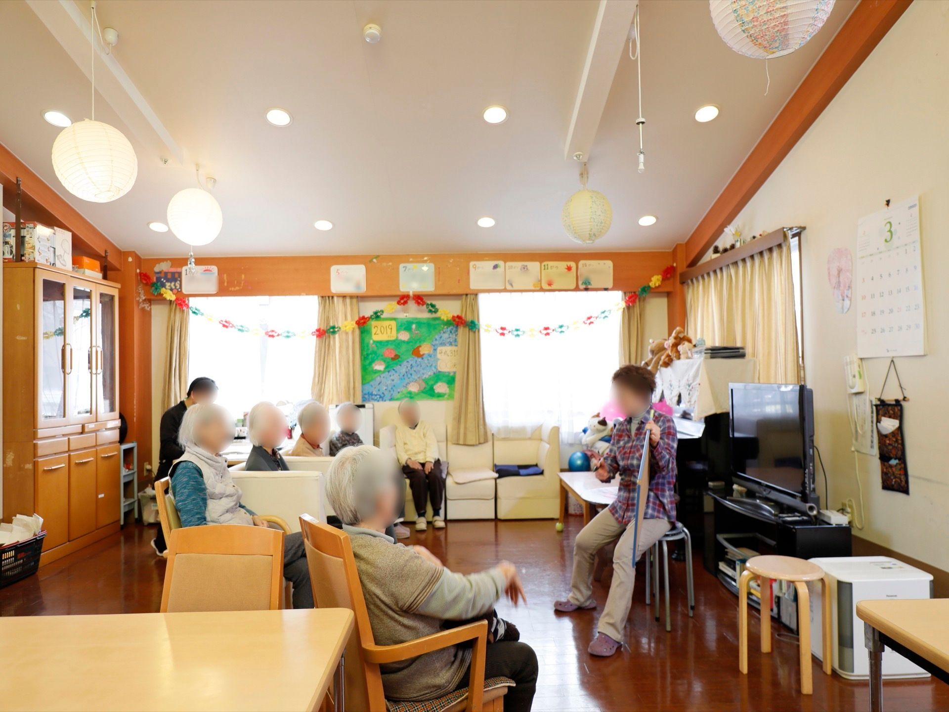 共有スペースに入居者様が集まって、椅子やソファに座っている。一人が前にある椅子に座って、ボードを使って話をしている。