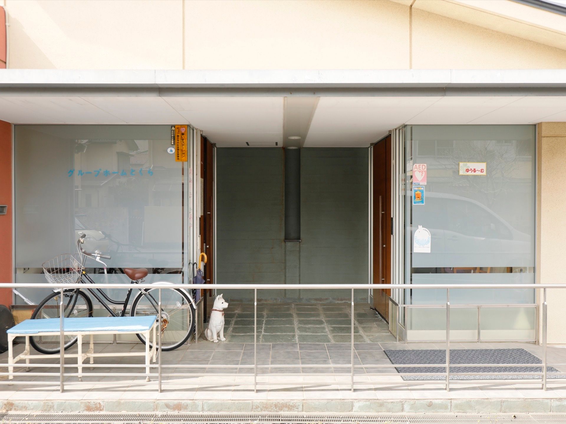 玄関は奥まった所にあり、左右に入口の扉がある。大きなひさしがあり、自転車が停められている。犬の置物がある。