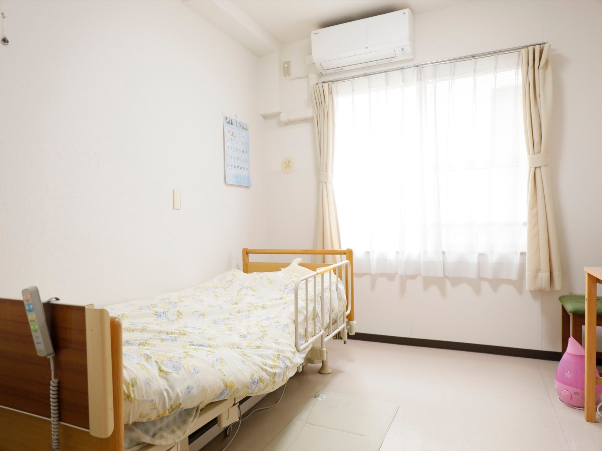 全面白色を基調とした明るい雰囲気の居室である。転落防止柵つきのベッドやエアコンが設置されている。加湿器が置かれている。