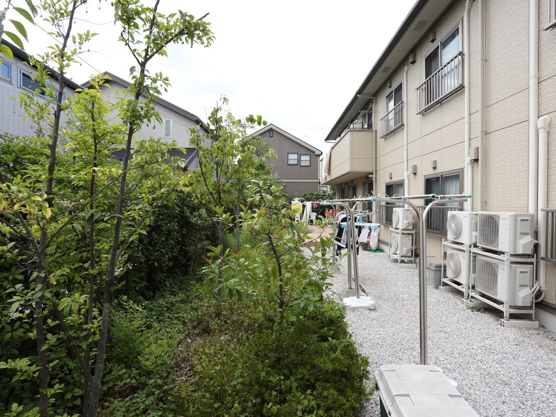 ２階建ての建物に沿って、砂利を敷いたスペースがある。物干し台が置かれ、洗濯物が干されている。脇には植物が茂っている。