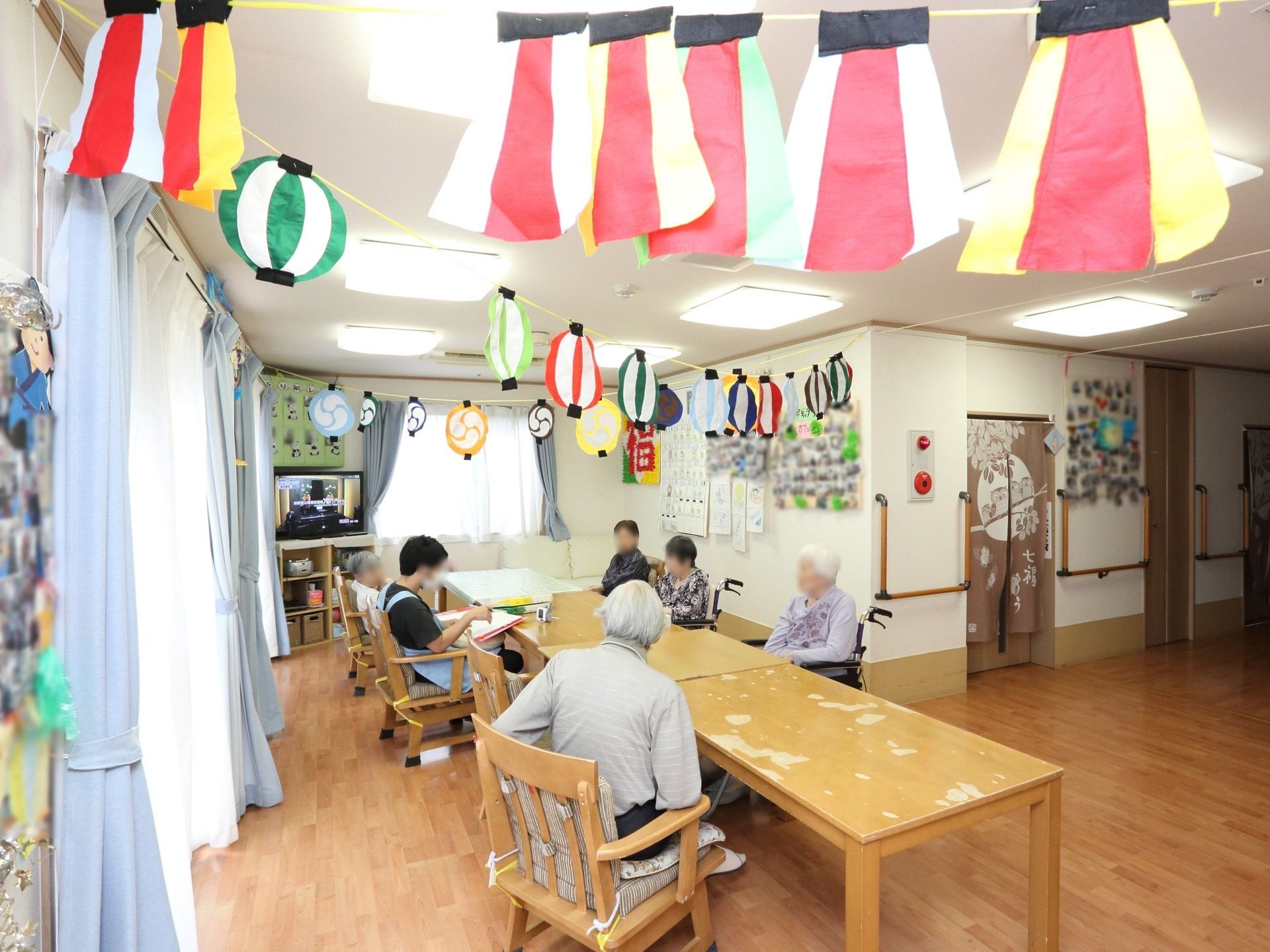 窓に囲まれた広く明るい談話室である。天井には手作りの提灯や和風の飾りつけがされている。隅にはテレビがあり、壁には飾りや写真が貼られている。