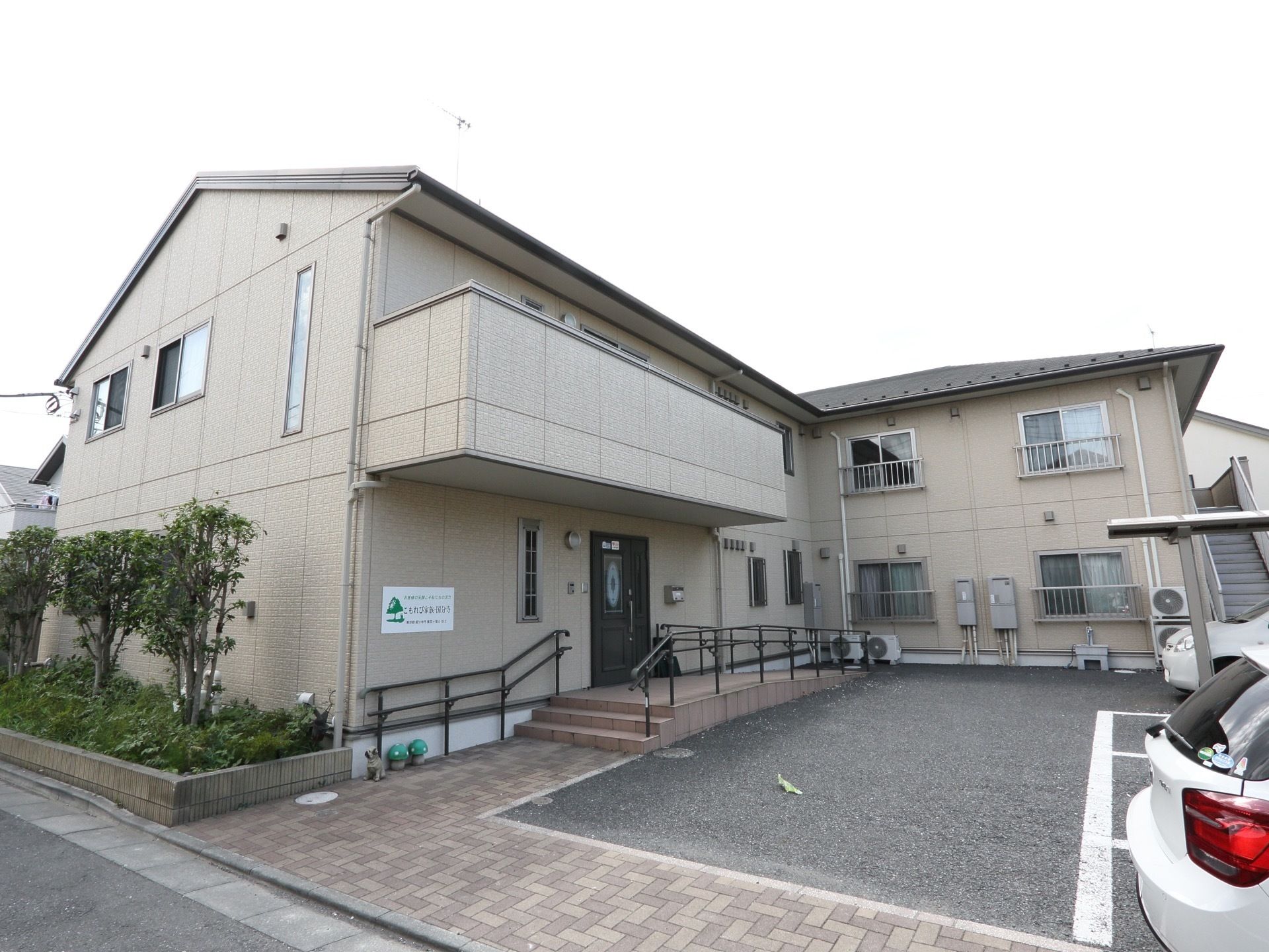 Ｌ字型の２階建ての建物の玄関先には、手すりのついたスロープと階段が設置されている。正面には駐車場がある。