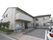 Ｌ字型の２階建ての建物の玄関先には、手すりのついたスロープと階段が設置されている。正面には駐車場がある。