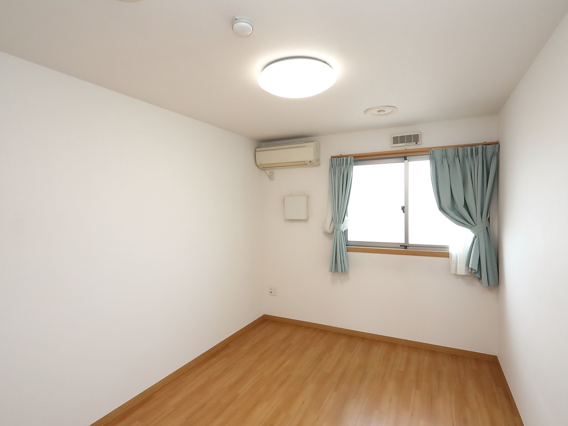 長方形の居室で、フローリングの床と白い壁でできている。右奥に窓があり、青いカーテンが下げられている。