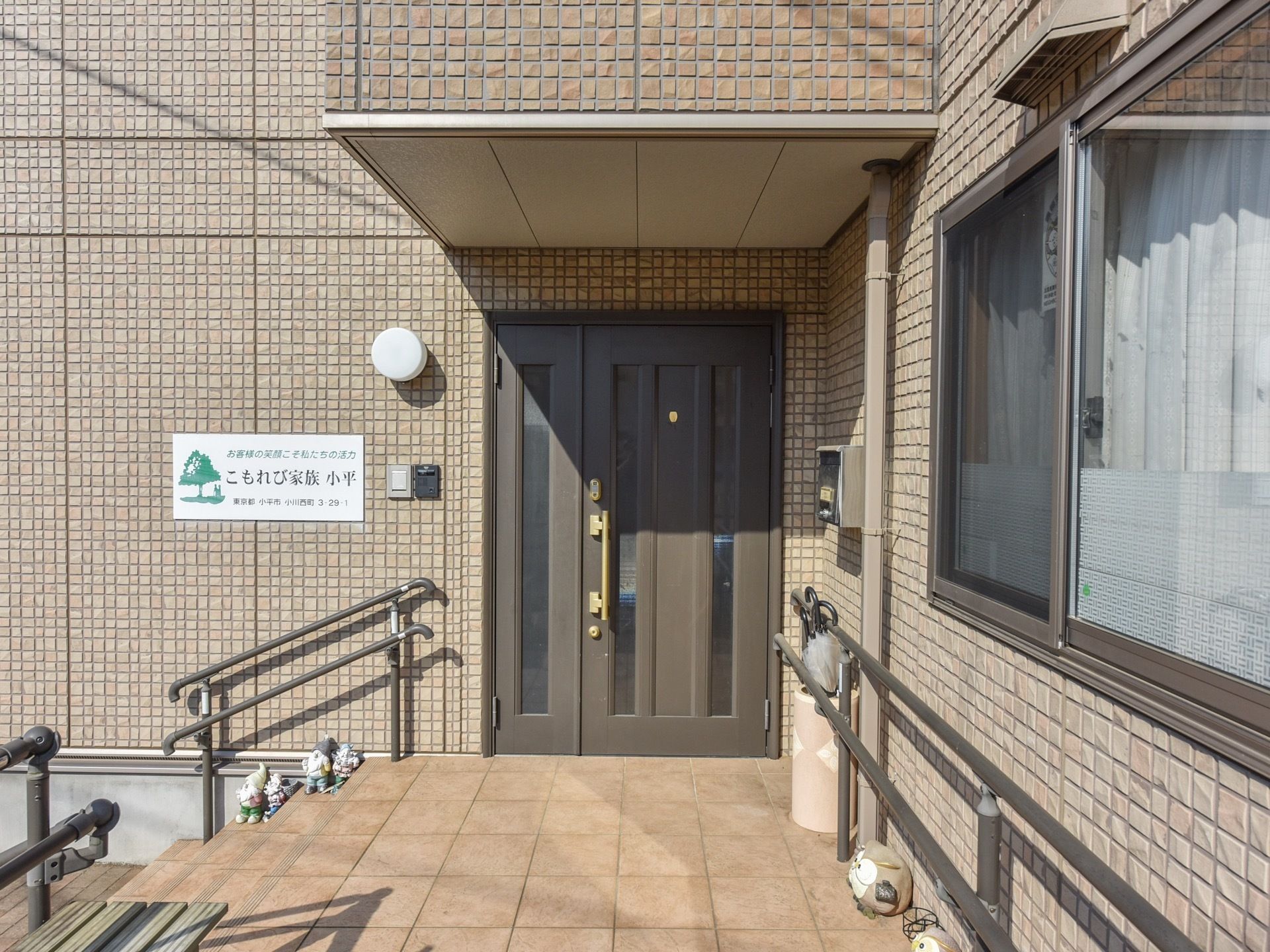 玄関ドア横の淡い赤茶色の壁に、施設名の入ったプレート板が取りつけてある。入口までの通路には手すりが設置されている。