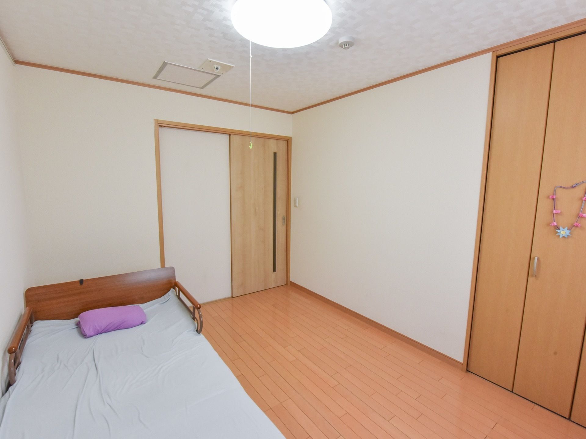 すっきりとしたフローリング床の部屋にベッドが置かれている。入口のドアは引き戸式である。ベッドの向い側の壁に収納庫が設置されている。