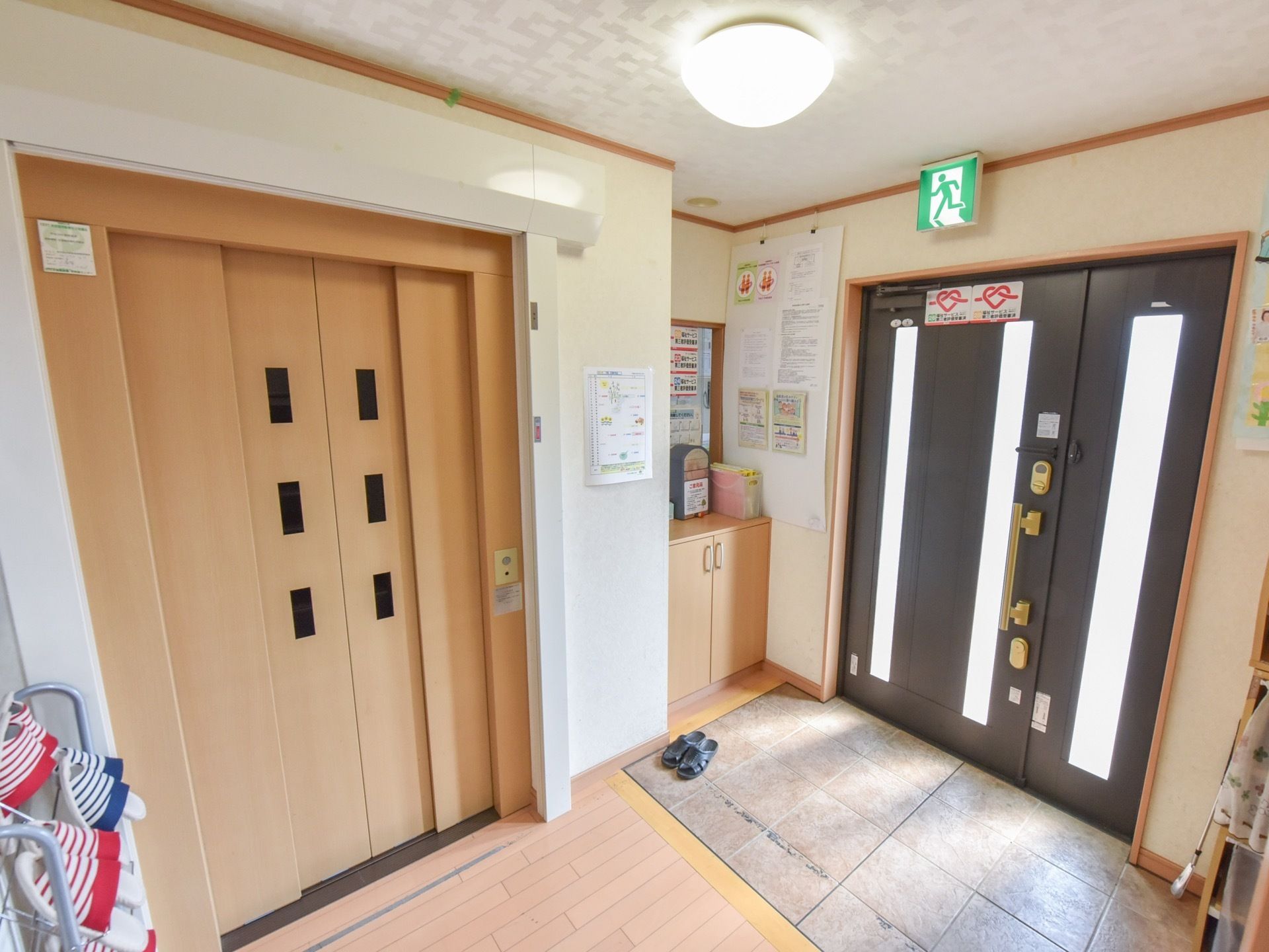 玄関ドアのガラスを通して、自然光が差し込んでいる。玄関横には下駄箱が設置されている。すぐ横には茶色のドアのエレベーターがある。
