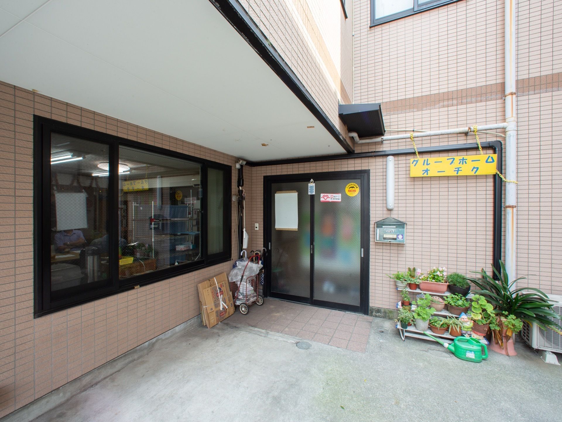 玄関扉はすりガラスをはめ込んだ引き戸となっている。雨どいを利用して、施設名が書かれた黄色の看板を設置している。