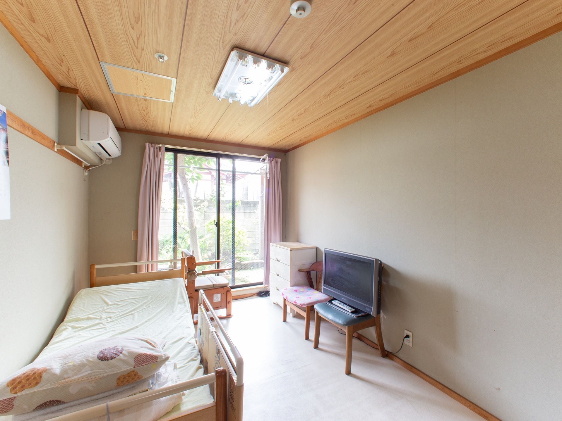 居室には、エアコンと介護ベッドが備え付けられている。白い収納ボックスの横に、椅子が２脚あり、うち１脚の椅子にはテレビが置かれている。