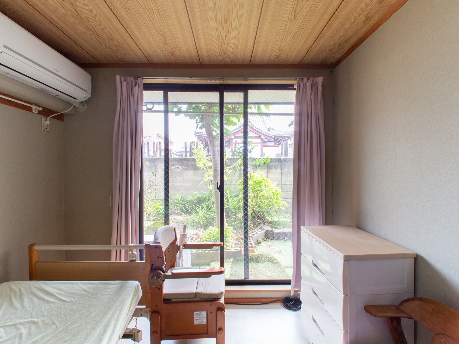 エアコンと介護ベッドが備え付けられた居室である。ベッド脇にはポータブルトイレが置かれている。サッシ越しに施設の庭が見える。