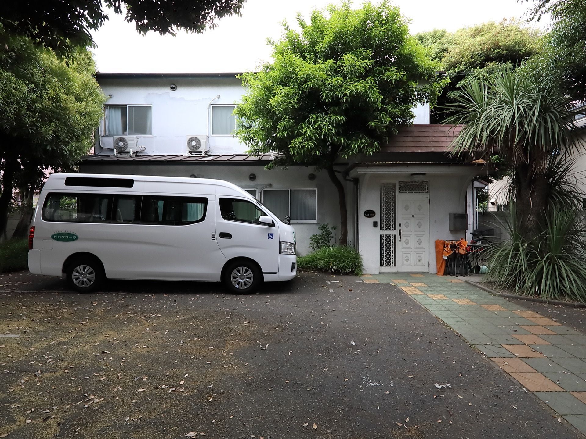 入口の両横に大きな木が植えられた2階建て施設である。地道の駐車スペースが設けられ、施設専用者が停車している。