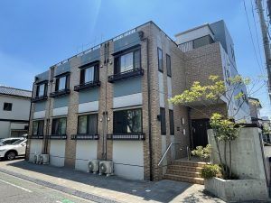 ２階建ての建物は日当たり良好でレンガ調の外壁。道路に面していて玄関には手すり付きの階段がある。
