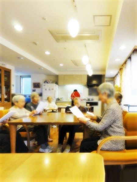 キッチンでスタッフが作業をしていて、手前のダイニングテーブルには入居者が集まっている。
