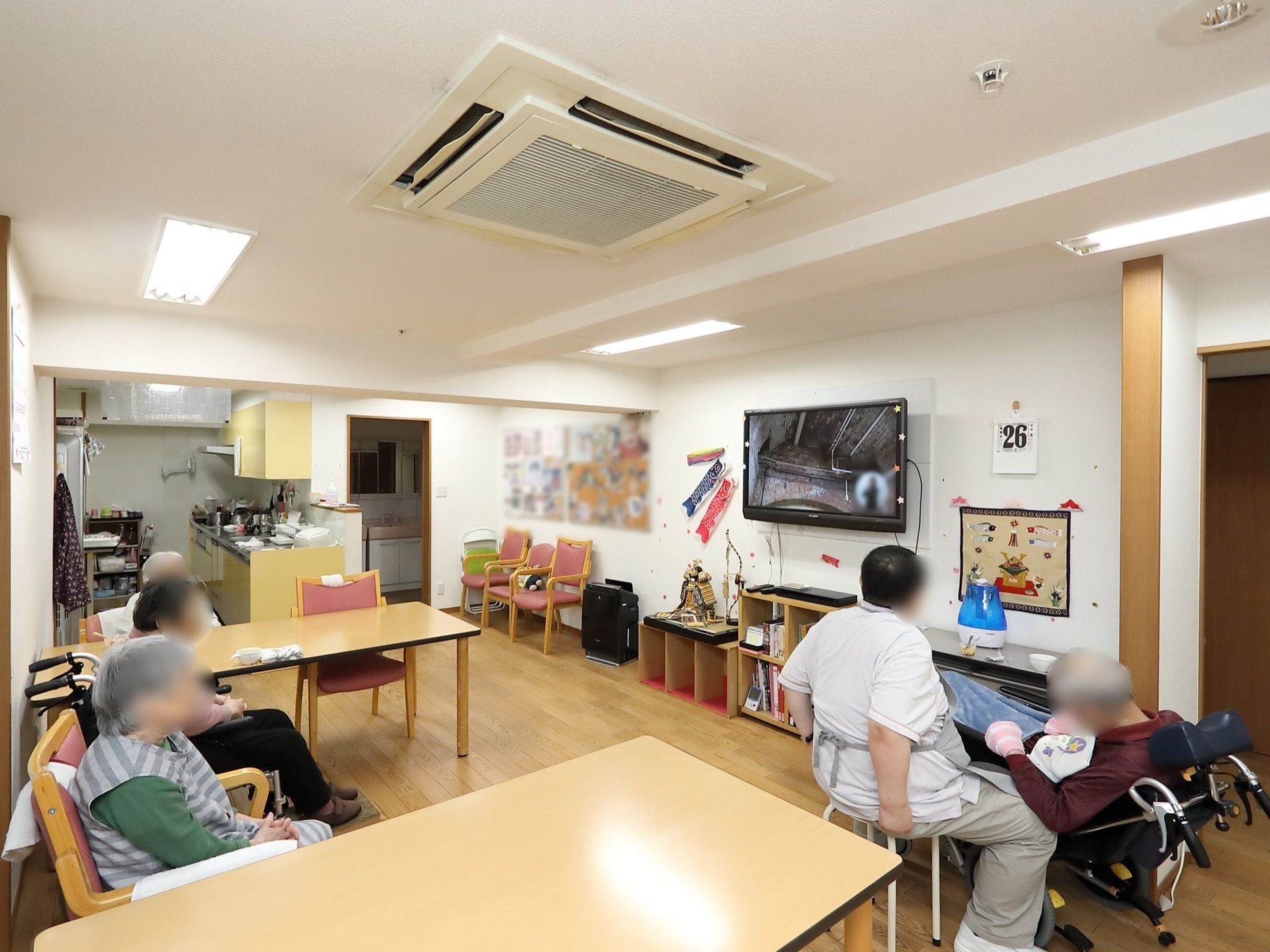 共有スペースには、壁掛けの大型テレビが設置されている。壁には季節感のある掲示物やカレンダーが貼られている。
