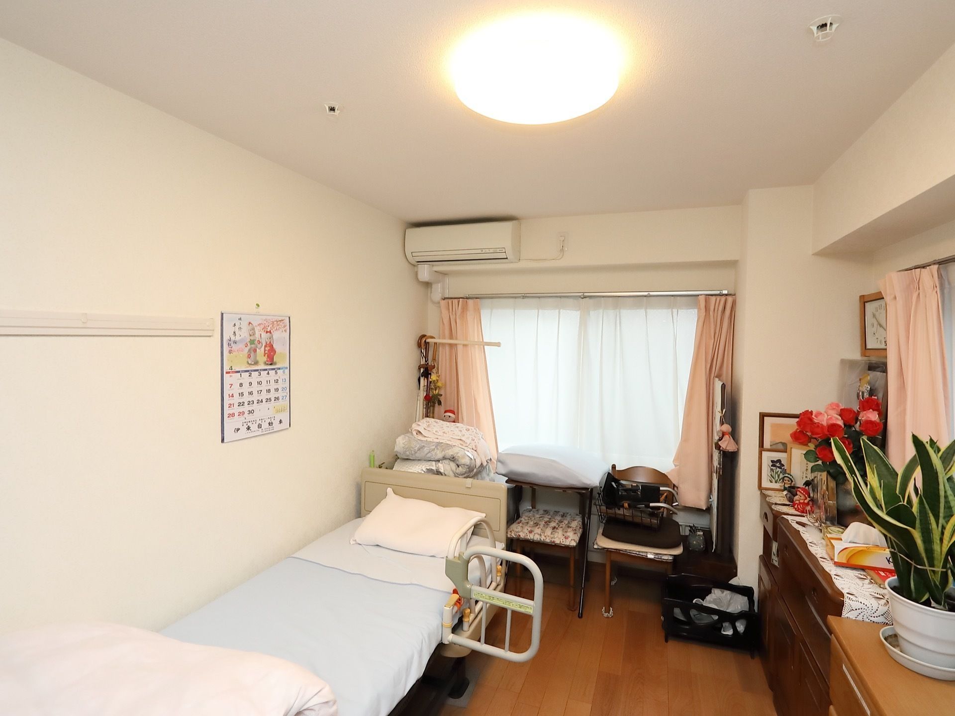窓が２箇所設置され、暖色系のカーテンがかけられている。また、壁際にベッドがあり、たんすや植物も置かれている。