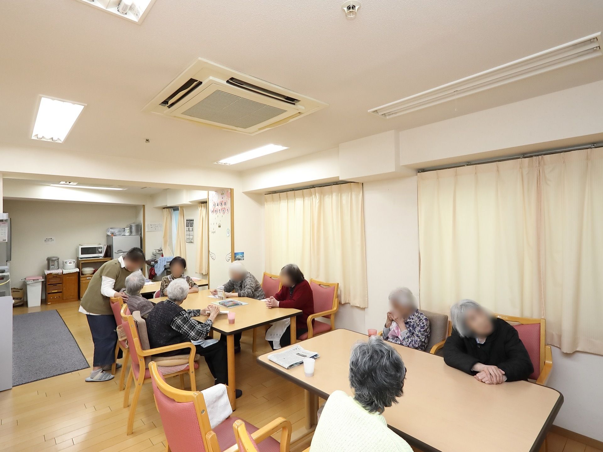 四角い４人がけのテーブルが置かれており、入居者様がお茶を飲みながら歓談している様子である。天井には埋め込み式のエアコンがある。
