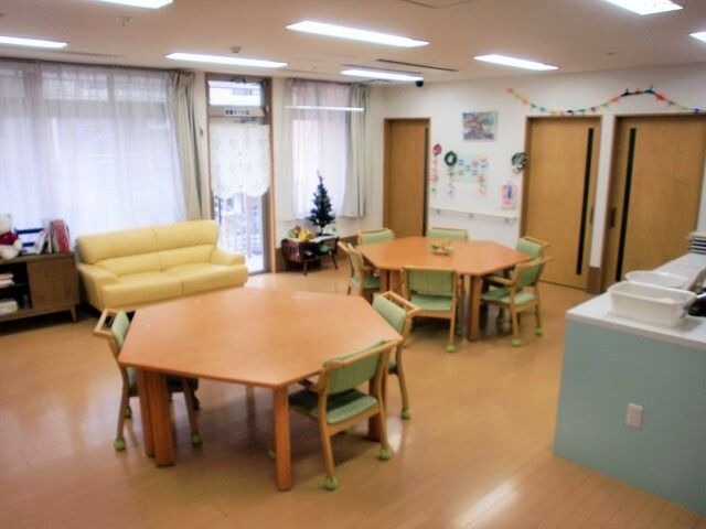 小さなクリスマスツリーが飾られたリビングダイニング。六角形のテーブルセットが二組置かれ、窓際には黄色いソファがあり、壁には手すりがある。