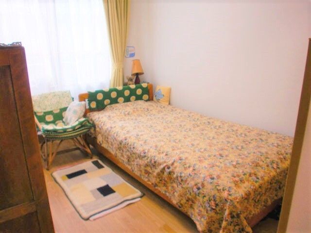 カーテンとレースカーテンが付いた大きな窓があるフローリングの居室にはベッドがあり、寝具がセットされている。椅子やマットもあり、個性が出ている。