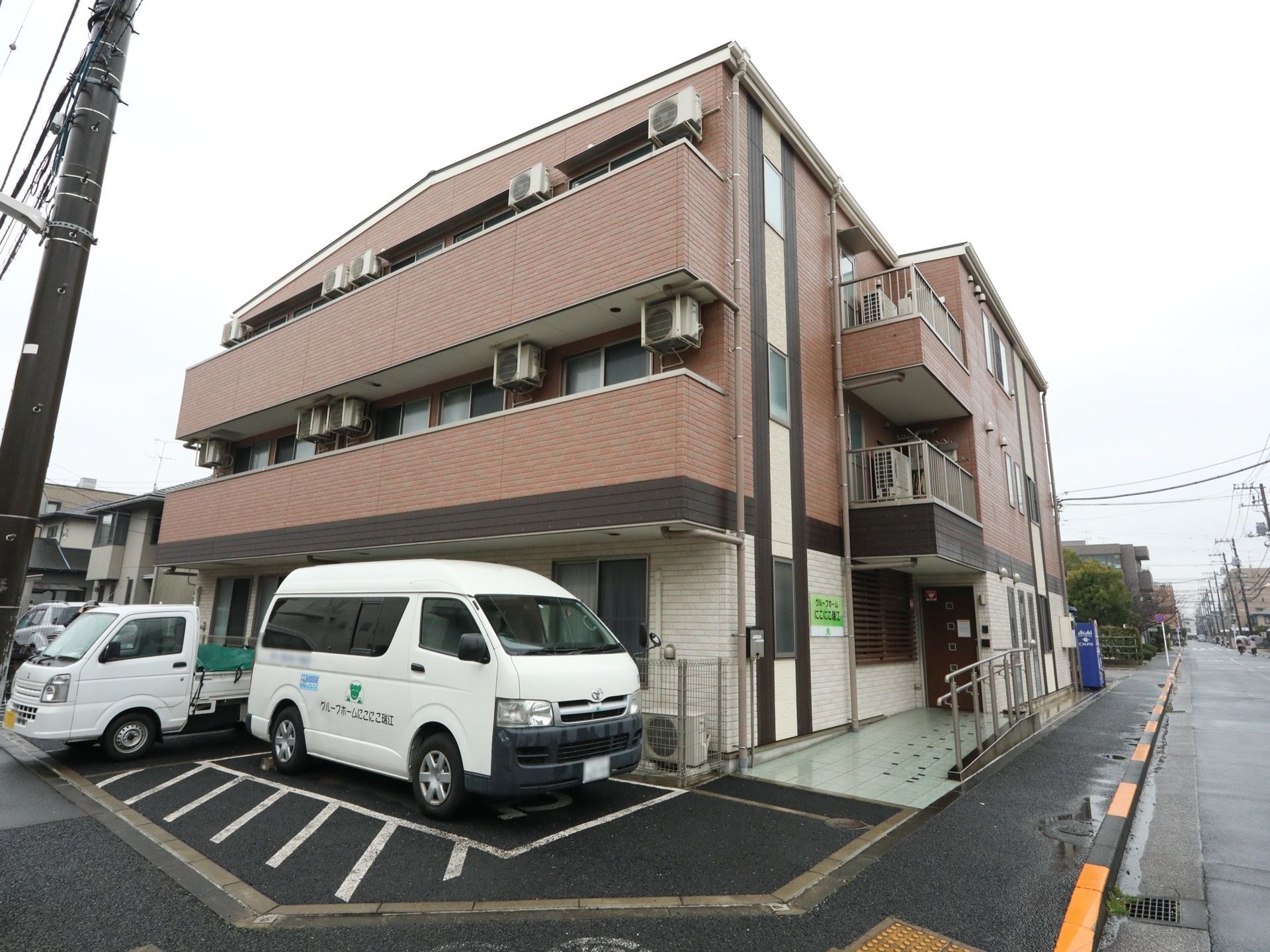 施設は３階建てで、外壁は茶色となっている。各階にベランダが付いている。建物の横に駐車スペースがあり、車が停まっている。