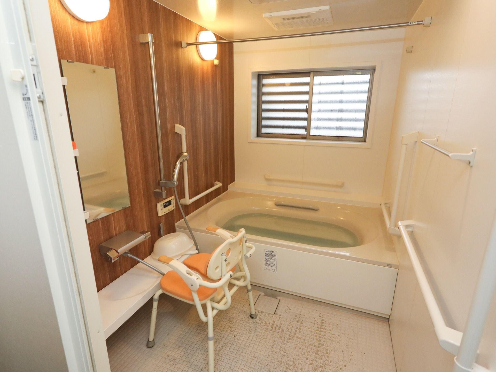 大きな窓の付いた浴室である。左側の壁は木目調で、ミラーやシャワーが付いている。洗い場や浴槽脇の壁に手すりが設置されている。