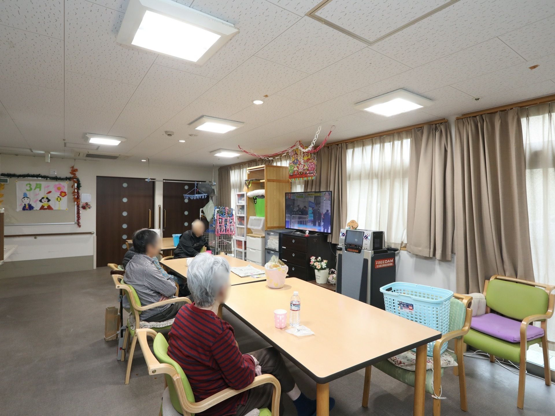 共有スペースの中央に、テーブルと椅子がある。。窓際にテレビが設置されていて、数名の入居者様が座って観ている。
