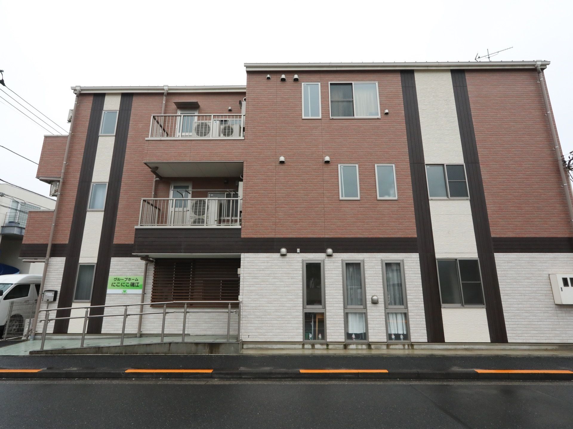 公道の脇に建てられた施設である。１階外壁は白で２、３階は茶色系となっている。左手より玄関に向かって、スロープの設けられた通路が伸びている。