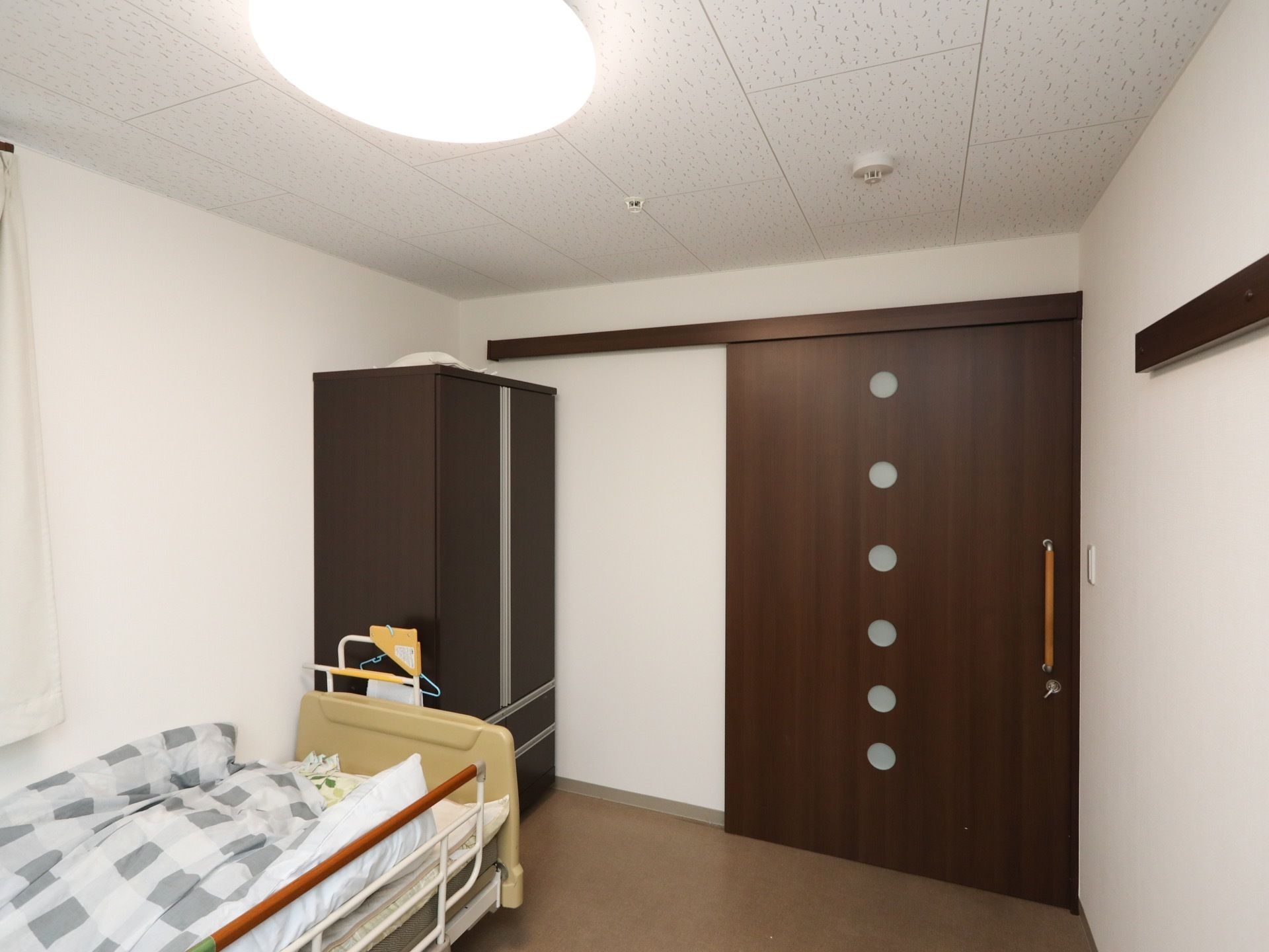 居室の入口ドアは、引き戸式である。転落防止柵の付いたベッドが置かれている。その横に茶色の収納タンスがある。