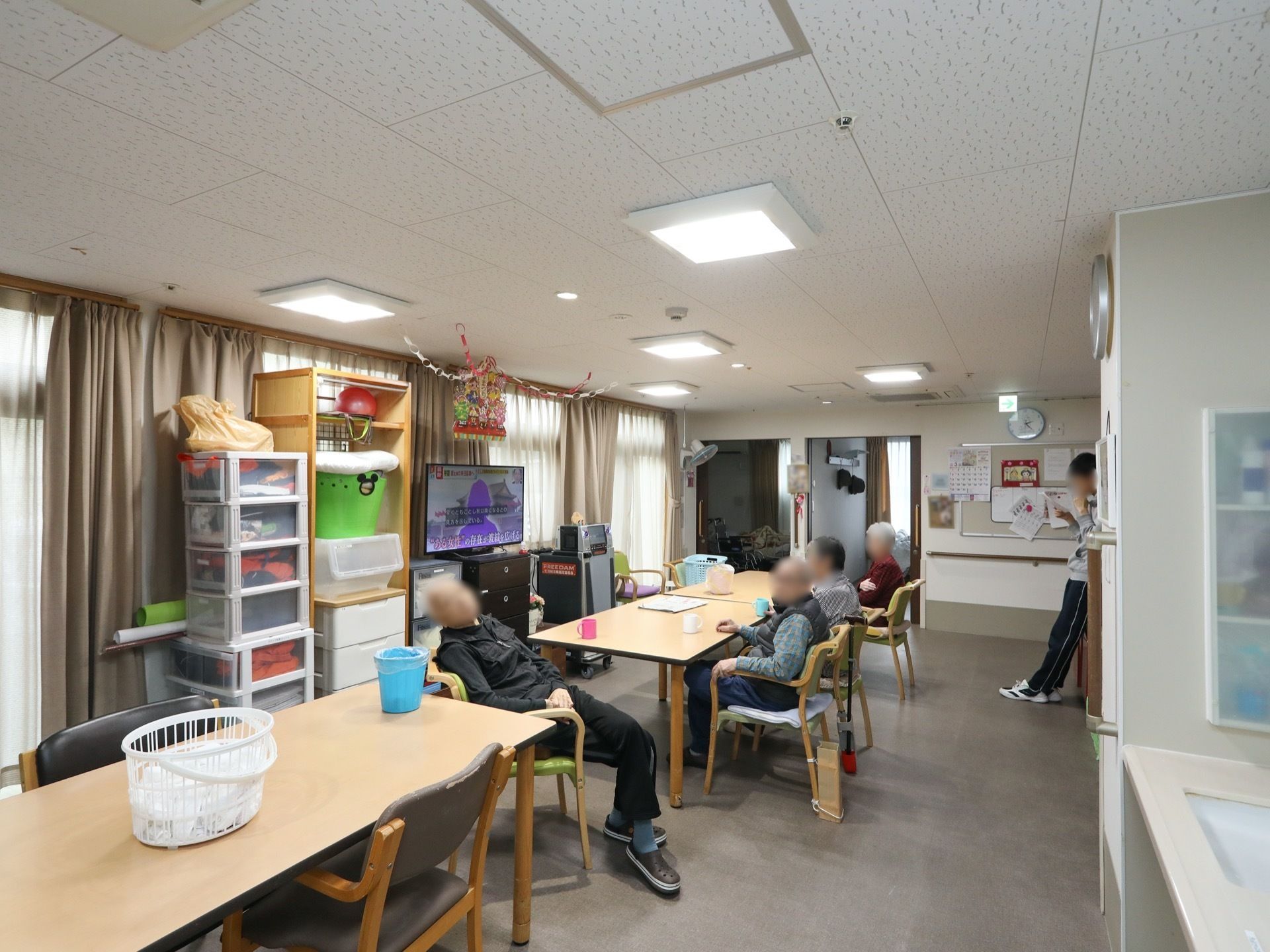 奥行きのある共有スペース。中央には、テーブルと椅子が縦に並べられている。窓にはカーテンが掛かり、その前に収納ケースやテレビが置かれている。
