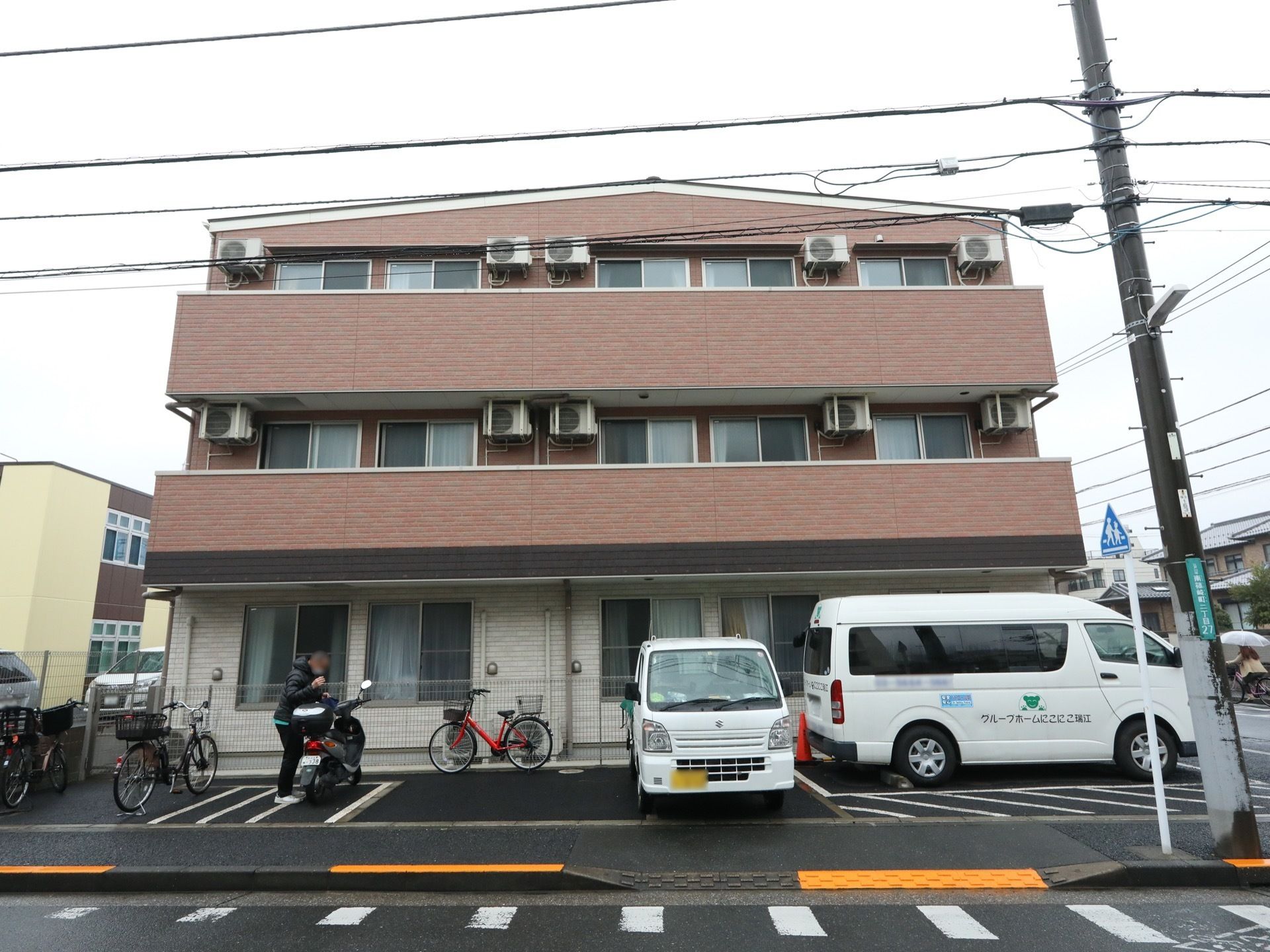 施設の建物側面の様子である。建物脇に駐車スペースが設けられている。車やバイク、自転車が停めてある。公道との間には、歩道が設けられている。