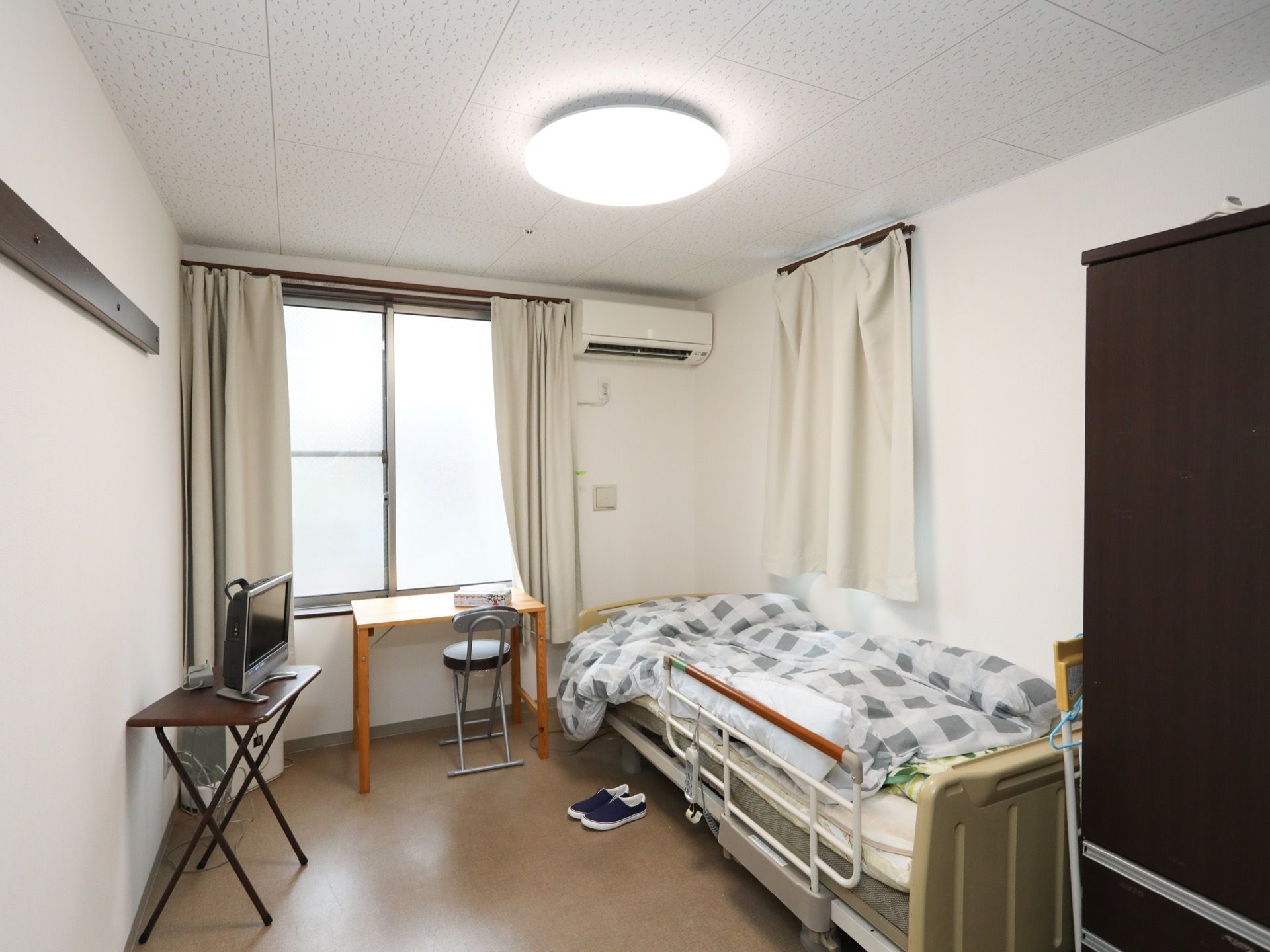 腰高の窓からの日差しで明るい居室である。窓下に、机と椅子がある。。ベッドの向い側の台の上にテレビがある。