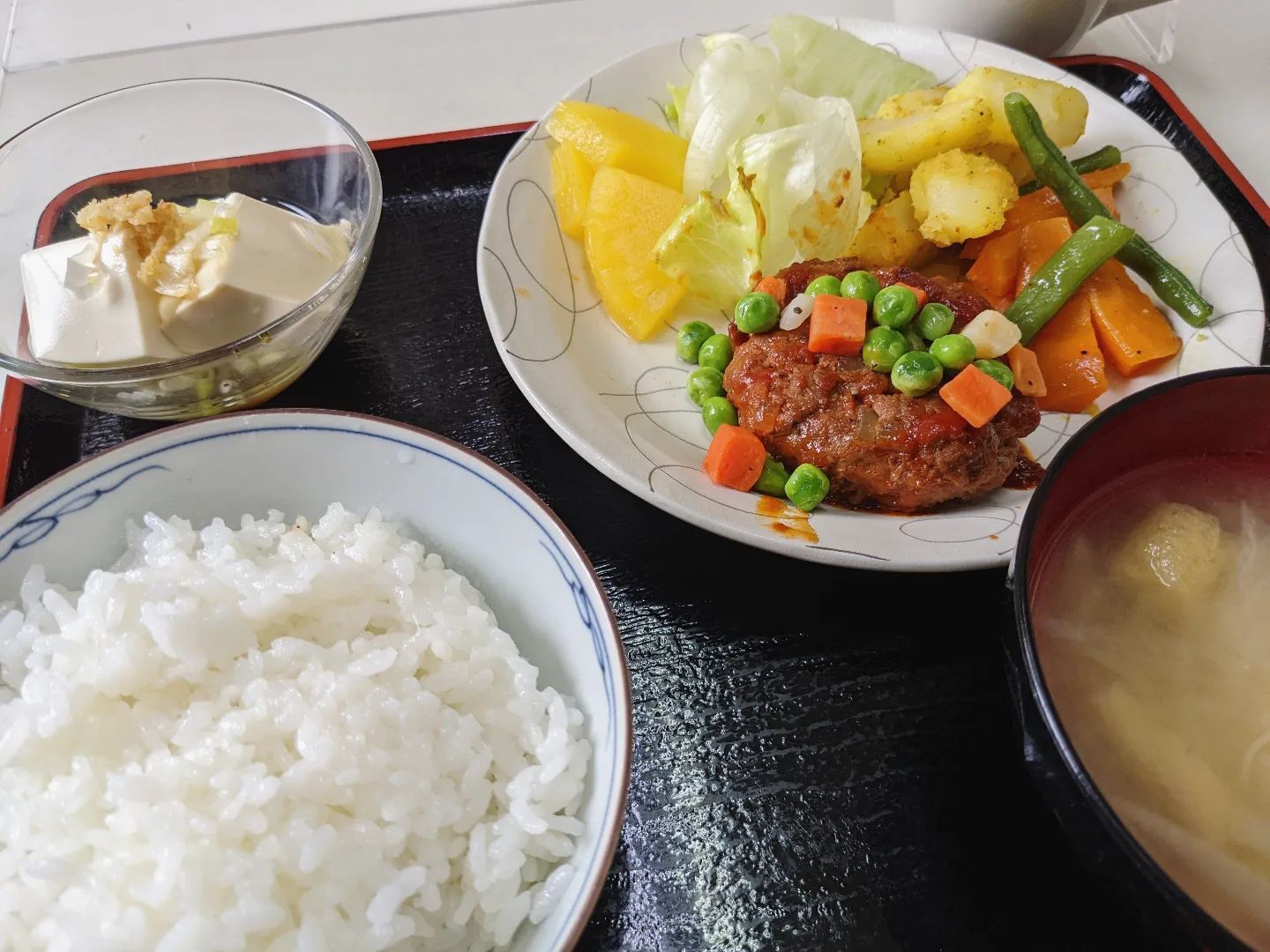 白いご飯、みそ汁、豆腐、野菜が添えられたハンバーグなどが並んでいる黒いトレー。
