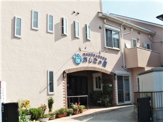 ２階建てのベージュの建物はモダンな雰囲気で、玄関周辺には花や緑が並んでいる。
