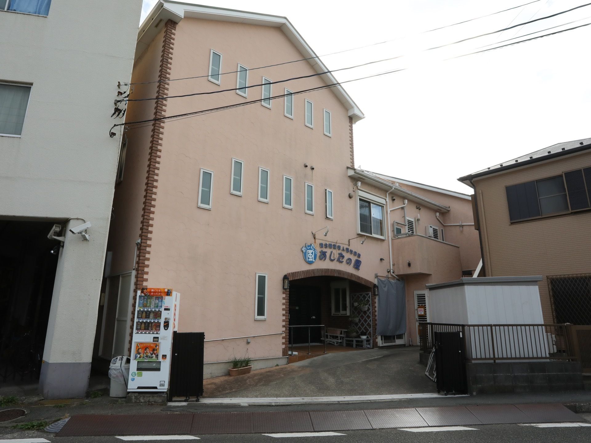 ピンクを基調とした、３階建ての三角屋根の施設。外壁には施設名が掲示されており、施設の前には自動販売機が設置されている。